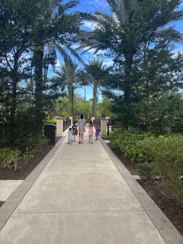 A tan concrete walkway lined with palm trees. A woman and 4 young children are walking off to the distance. 