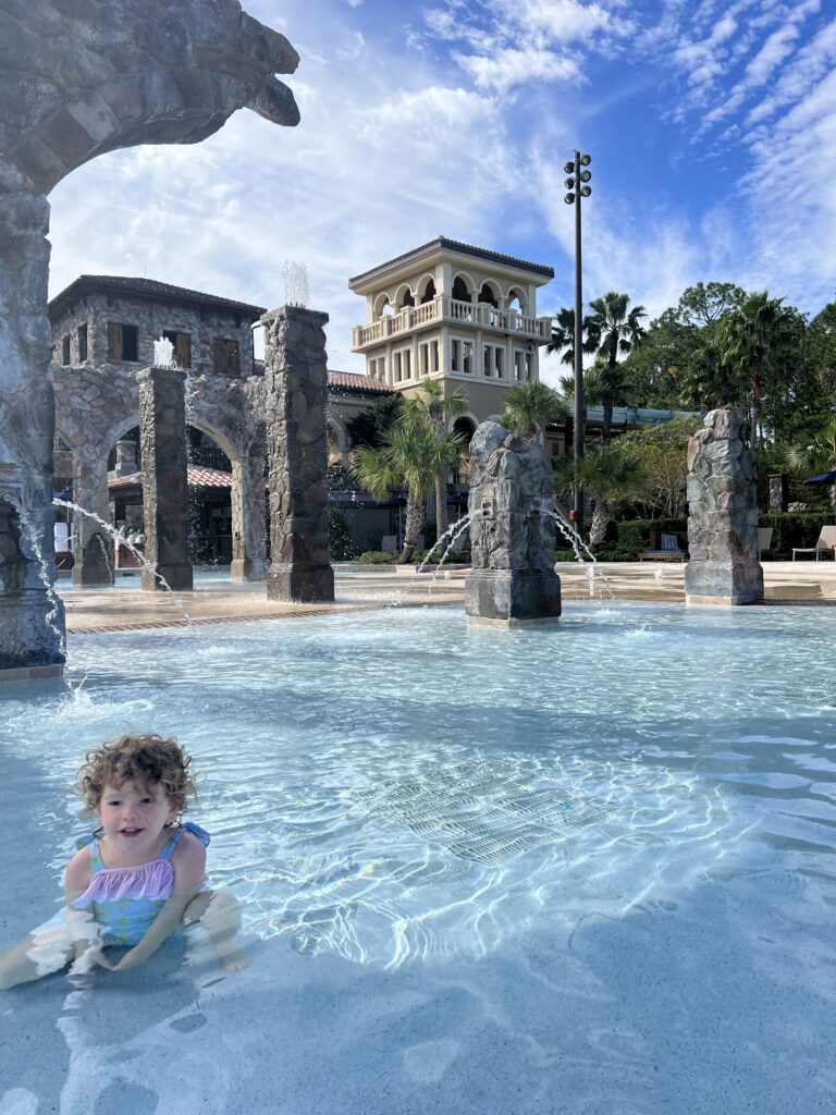 A young girl in a pink and blue bathing suit is in a kids pool with water. There are fountains in the background and blue skies. 