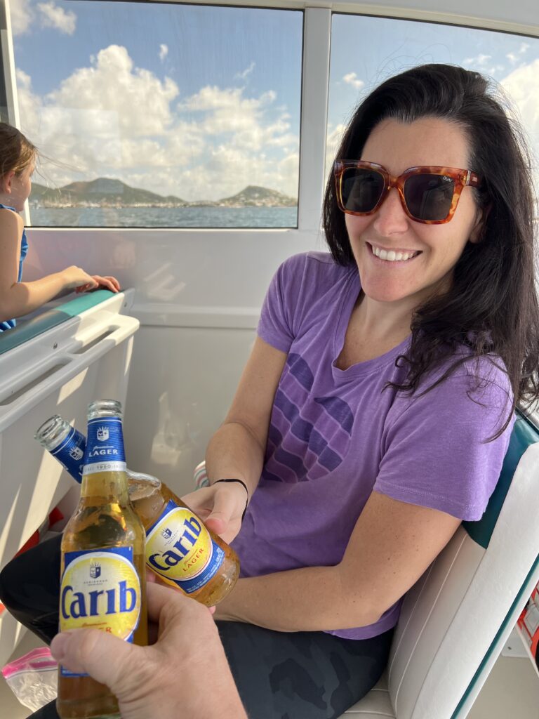 A woman in a purple shirt with brown sunglasses drinking an Anguillan beer with water and mountains in the background. She is cheersing the photographer. 