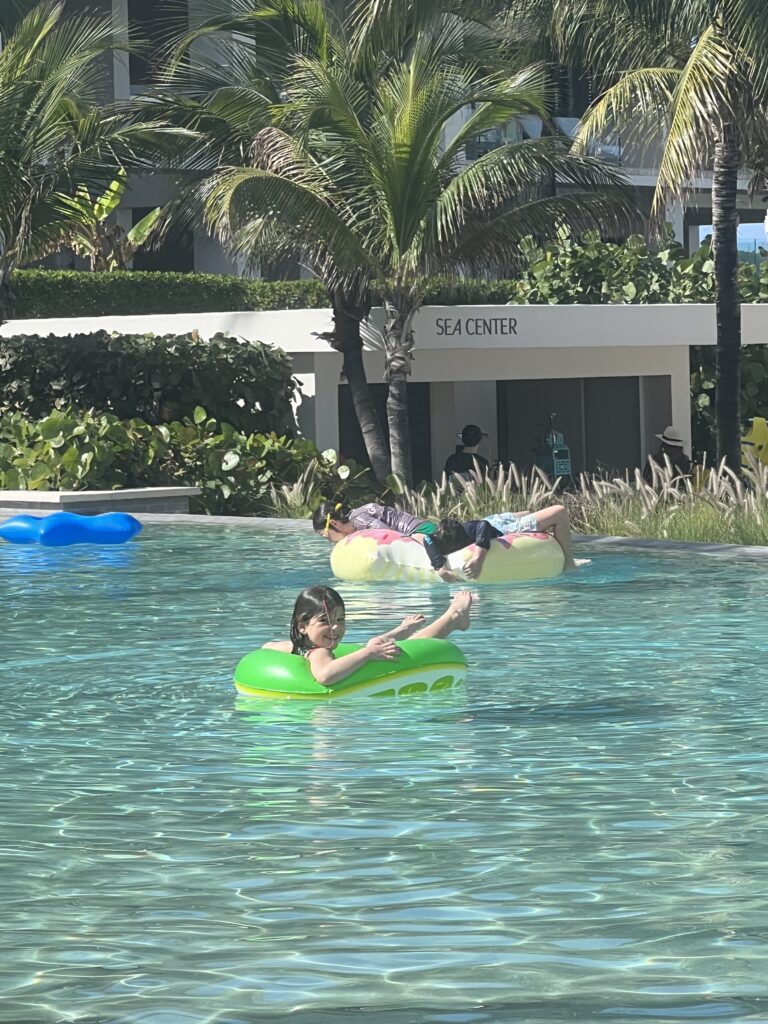 A young girl in a green inner-tube is floating in a shallow pool. A building is in the distance with palm trees. 