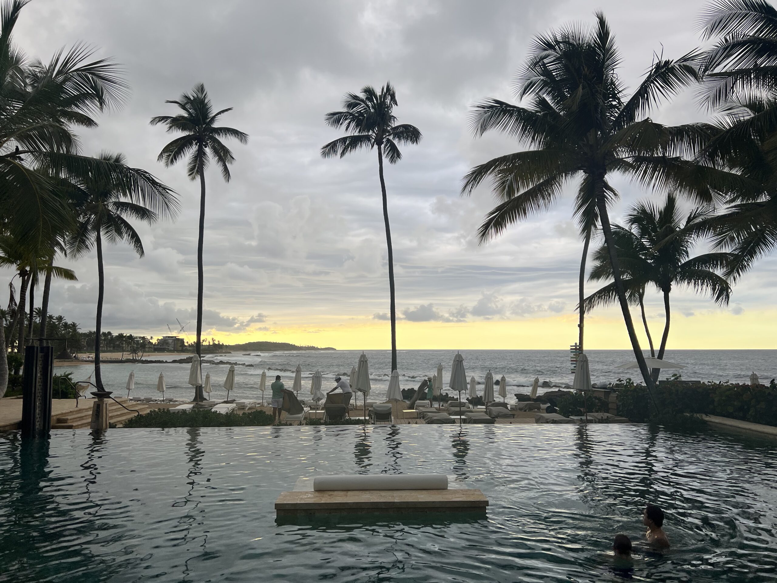 A pool in Puerto Rico overlooking a sunset with palm trees and beach chairs in the distance.