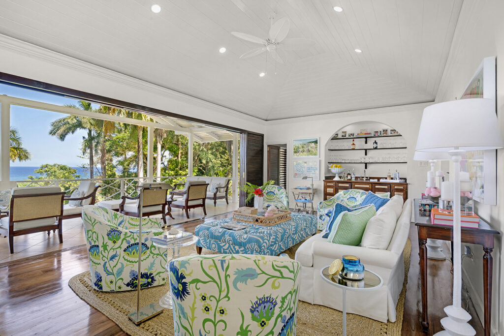 View of living room with colorful, blue and green fabrics. A bar is in the distance, as well as ocean views outside of the open doors. 