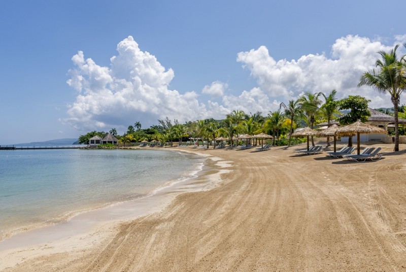Image of a sunny, empty beach. Lounge chairs under grassy umbrellas. 