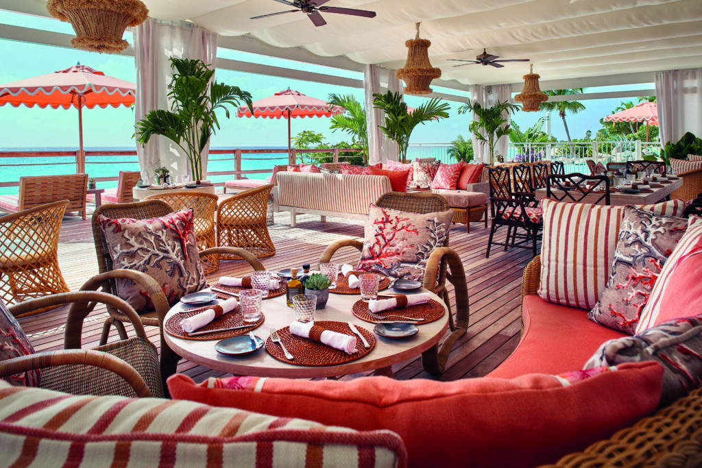 Coral pink and White textiles adorn woven wood furniture on an open air dining space that overlooks the sea. 