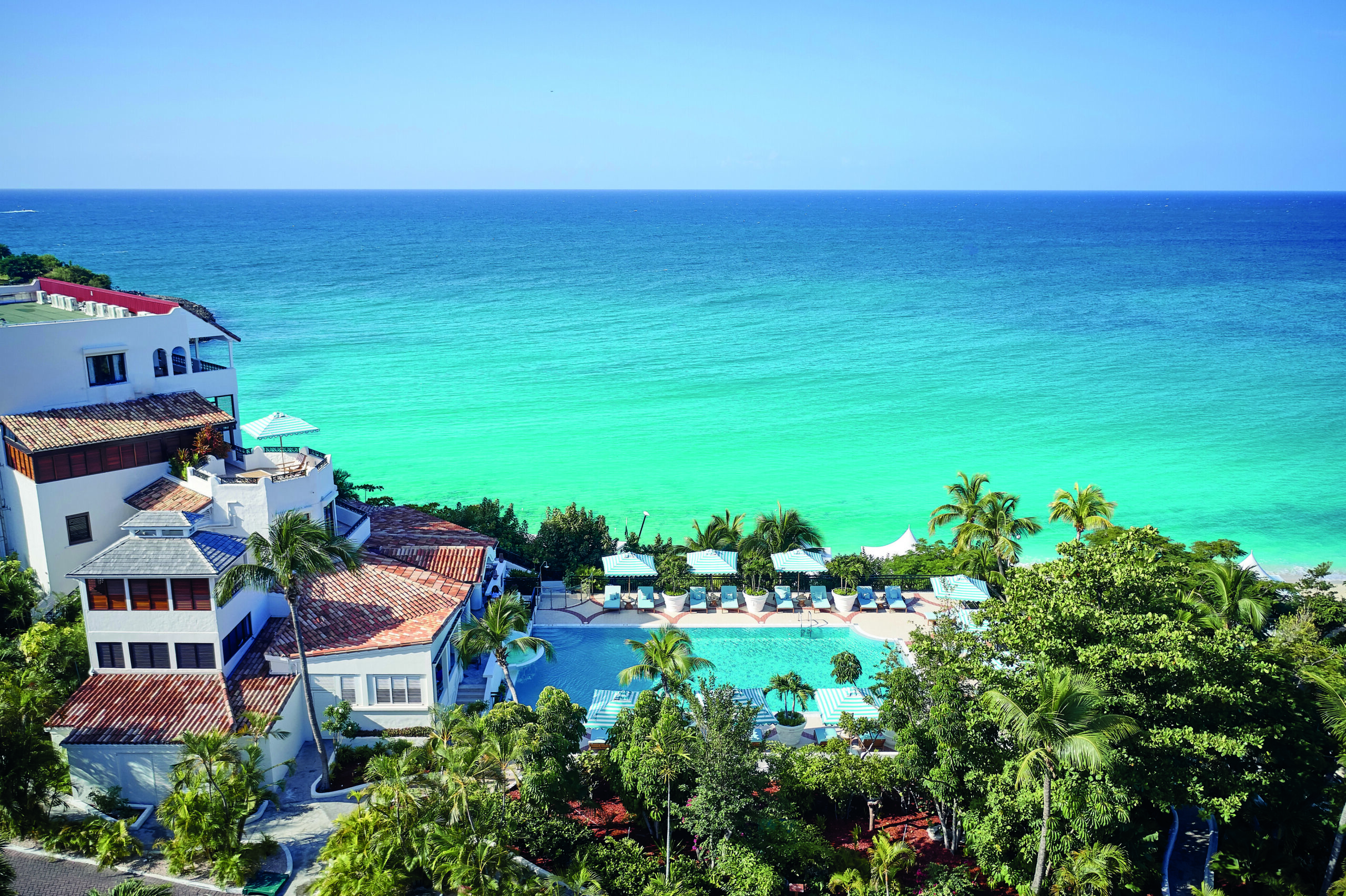 Aerial view of red roofed, white buildings and a pool (bottom Right) with a turquoise ocean in the distance