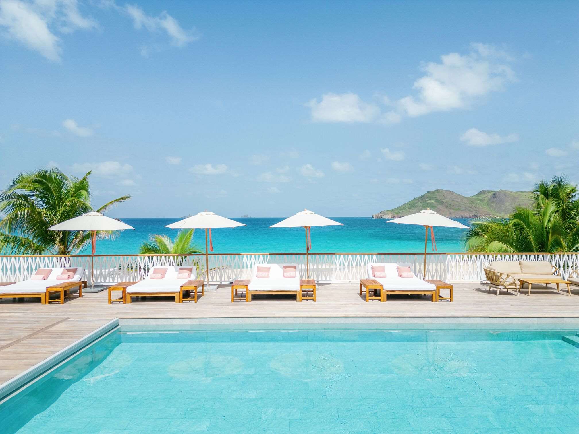 Pool with large lounge chairs with umbrellas in the background. Sea is visible in the distance.