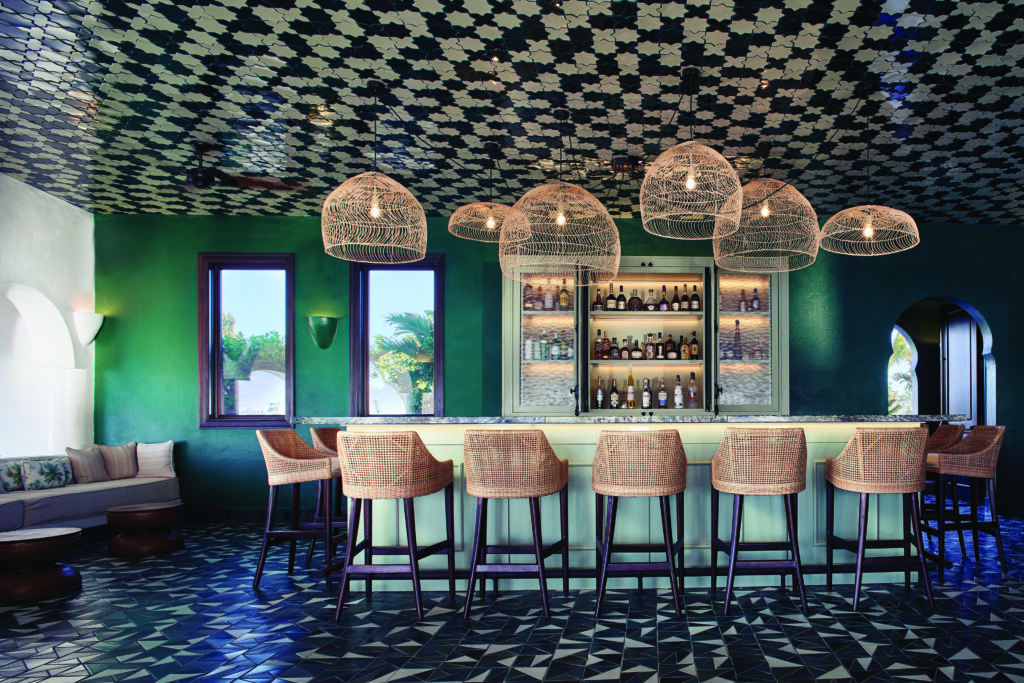 A green bar is surrounded by woven wood bar stools and woven wood pendants. The floor and ceiling are black and white moroccan tiles