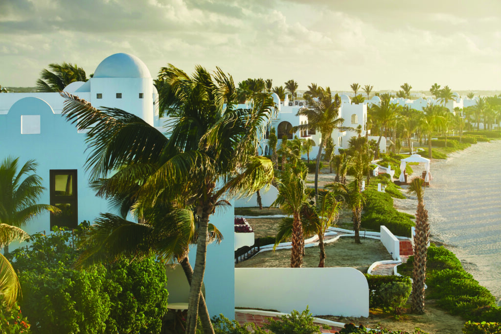 View of buildings surrounded by palm trees with the ocean to the right hand side. 