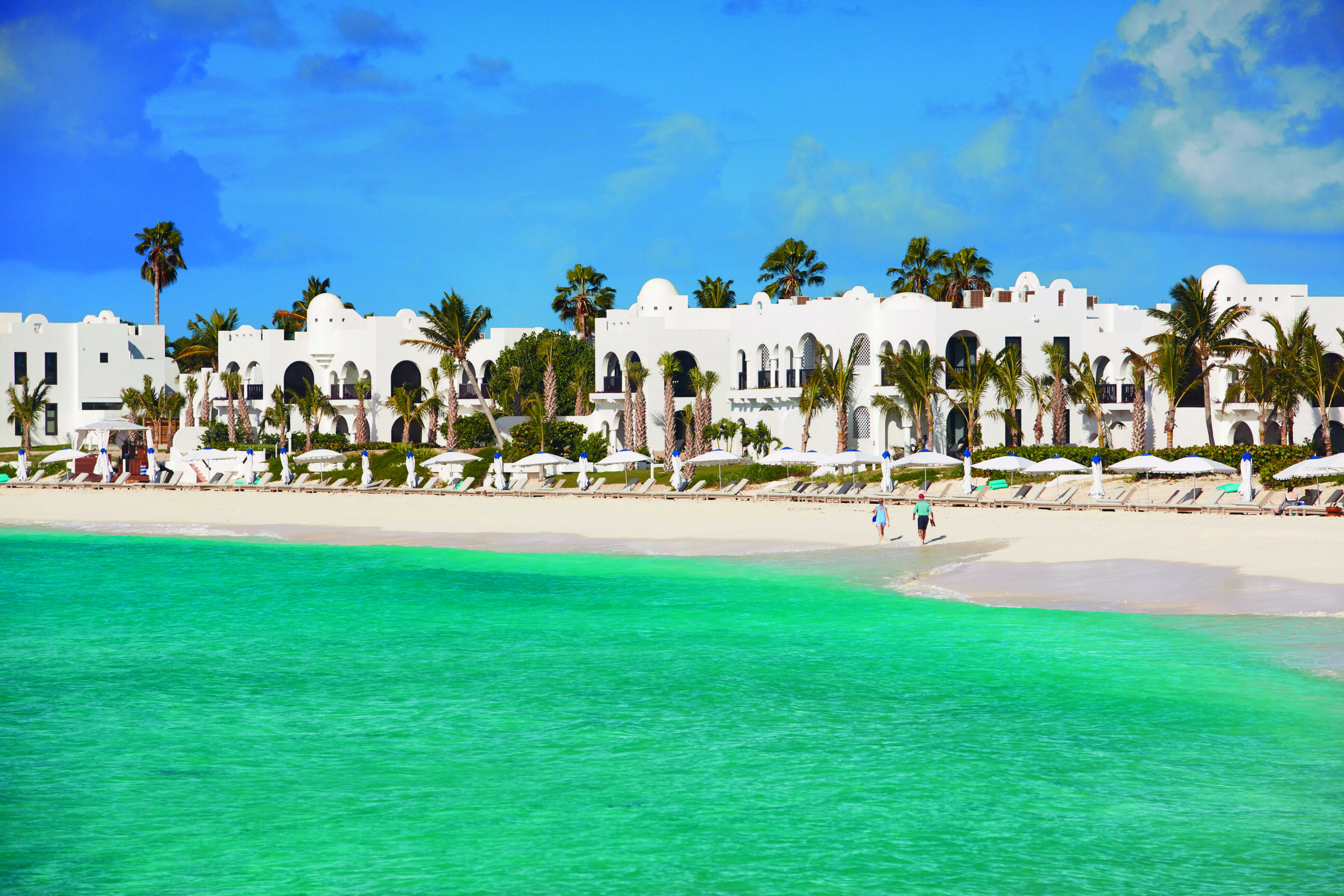 Turquoise and white sand beach with palm trees and white washed moroccan style buildings in the background.