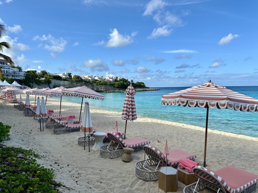 Coral pink and white lounge chairs with striped umbrellas spread along a white sandy beach with turquoise water on Baie Lounge Beach.